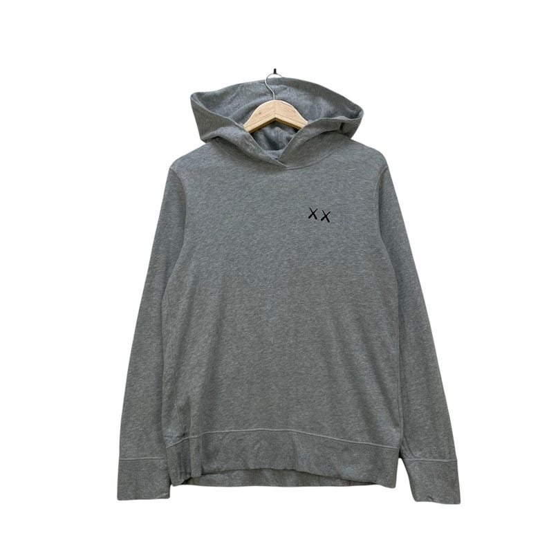 Peut inclure: Un sweat &agrave; capuche gris avec une capuche et des manches longues. Le sweat &agrave; capuche porte un petit logo noir "XX" sur la poitrine. Le v&ecirc;tement est suspendu &agrave; un cintre en bois.