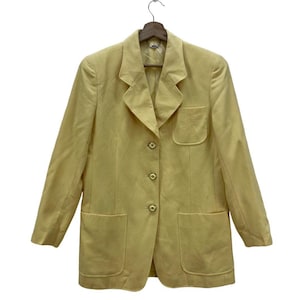 May include: A light yellow blazer with a notched collar, three buttons, and two front pockets. The blazer is hanging on a wooden hanger. The jacket has a pocket on the left side.