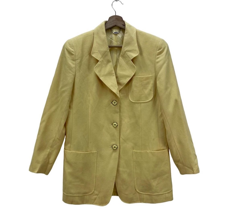 May include: A light yellow blazer with a notched collar, three buttons, and two front pockets. The blazer is hanging on a wooden hanger. The jacket has a pocket on the left side.