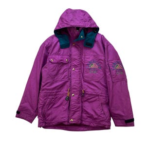 Peut inclure: Veste à capuche magenta avec un col vert d'eau et le texte "CABIN CLUB 1974" brodé sur la poitrine. La veste a un cordon de serrage, plusieurs poches et des boutons dorés. Les poignets sont noirs.