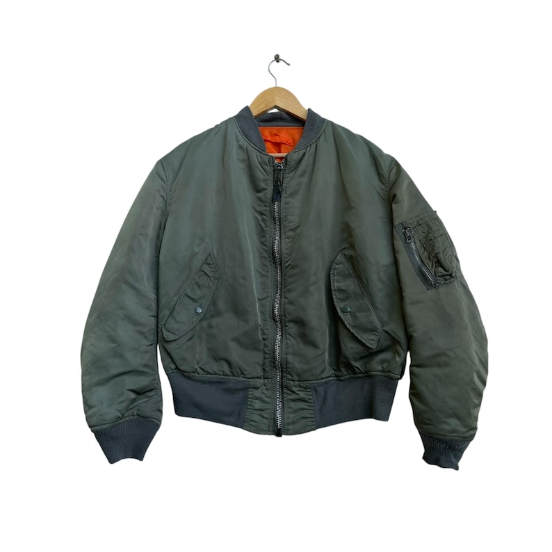 Puede incluir: Chaqueta bomber verde oliva con cuello, pu&ntilde;os y cintura grises. La chaqueta tiene cierre de cremallera, dos bolsillos delanteros y un bolsillo con cremallera en la manga izquierda. El forro interior es naranja. La chaqueta est&aacute; en una percha de madera.