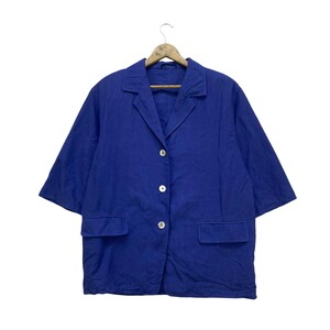 Puede incluir: Un blazer azul con tres botones blancos y dos bolsillos delanteros. El blazer tiene mangas cortas y cuello con solapas. Cuelga de una percha de madera.