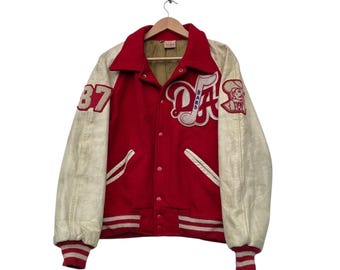 Op de afbeelding: Een vintage rode en crèmekleurige letterman-jas met het woord "BAND" en de letters "DA" in het wit. De jas heeft een rode body, crèmekleurige mouwen en het nummer "87" op de linkermouw.