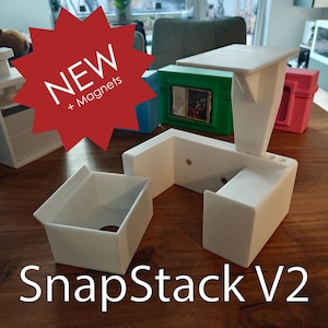 Könnte beinhalten: Weiße SnapStack V2-Organizer aus Kunststoff mit einem roten Stern-Grafik, die "NEW + Magnets" sagt. Im Hintergrund befinden sich mehrere farbige Boxen. Die Organizer stehen auf einem Holztisch.