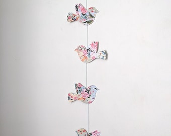 May include: A string of five paper birds with floral patterns hanging from a white string against a white wall. The birds are in various shades of blue, pink, and yellow.