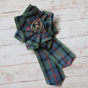 May include: A tartan ribbon brooch in blue, green, and red. The brooch has a silver-toned thistle emblem at its centre. The ribbon is arranged in a layered, flower-like design with a long, trailing tail, and green edging.