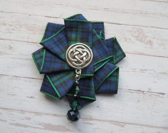May include: A blue and green plaid fabric brooch with a silver Celtic knot center and a dangling black bead accent. The brooch is made of folded ribbon and has a decorative design.