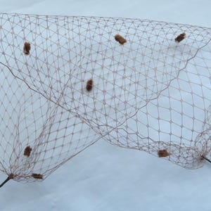 May include: A tan-coloured veil with a diamond-shaped mesh pattern. The veil is adorned with several small, brown, fuzzy embellishments. The veil is supported by two thin, dark brown stems, suggesting it is a headpiece.