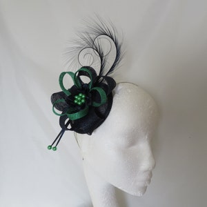 Peut inclure: Un fascinateur bleu marine et vert avec un motif floral et des plumes. Le couvre-chef présente une base bleu marine foncé avec des détails de ruban vert et un groupe de perles vertes au centre. Des plumes noires courbent vers le haut.