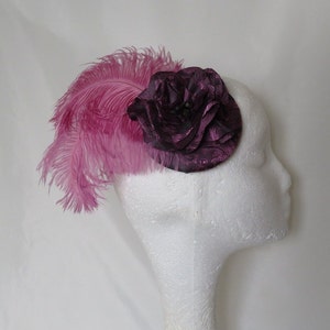 May include: A small, purple fabric flower with a pink feather accent. The flower is made of multiple layers of fabric and has a textured design. The feather is long and flowing.