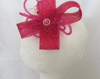Può includere: Un fascinator fucsia vivace caratterizzato da una base in tessuto a rete testurizzato, accenti di nastro ad anello e un ornamento circolare centrale con una perla rosa circondata da un anello di piccoli cristalli chiari, creando un aspetto floreale.