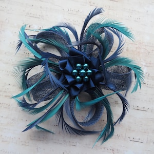 May include: A navy and teal feather fascinator with a central cluster of teal beads. The accessory features a dark blue satin bow and is accented with dark blue netting. The feathers are arranged in a radial pattern.