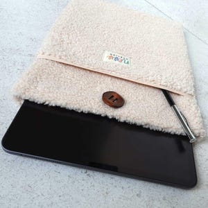 Op de afbeelding: Een beige, pluche tablet hoes met een houten knoopsluiting en een pennenhouder. De hoes heeft een kleurrijk "Nebula" label dat aan de voorkant is genaaid.