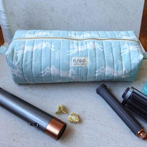 Può includere: Un beauty case trapuntato azzurro con un motivo a conigli bianchi e una cerniera dorata. La borsa ha un'etichetta con la scritta "neve atelier". Un asciugacapelli e strumenti per lo styling sono accanto.
