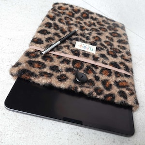 Może przedstawiać: Brązowo-czarna futerał na tablet z nadrukiem lamparta, różową wstążką i czarnym guzikiem. Futerał wykonany jest z miękkiego, puszystego materiału. Na futerału widoczny jest napis "nyrovo".