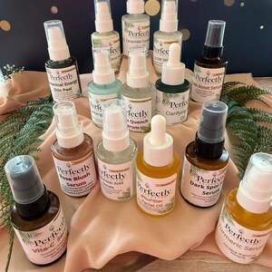 May include: A collection of skincare products in small glass bottles with white and black labels. The bottles are arranged on a surface with greenery and a dark blue background. The labels include text such as "Rose Blush Serum" and "Turmeric Serum".