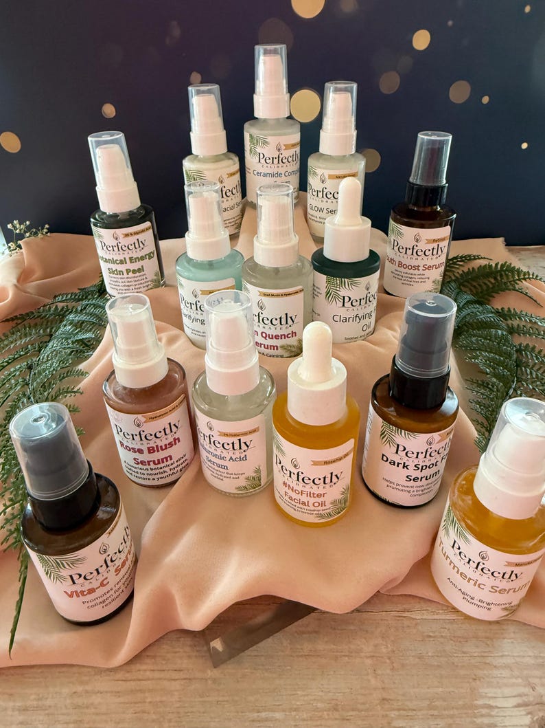 May include: A collection of skincare products in small glass bottles with white and black labels. The bottles are arranged on a surface with greenery and a dark blue background. The labels include text such as "Rose Blush Serum" and "Turmeric Serum".