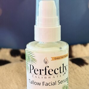 May include: A white glass bottle of Perfectly Calibrated Tallow Facial Serum with a pump dispenser. The label includes text and a palm leaf design. The product is on a clear, rectangular stand. The background is a blurred blue and brown.