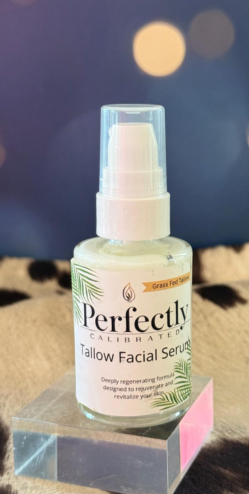 May include: A white glass bottle of Perfectly Calibrated Tallow Facial Serum with a pump dispenser. The label includes text and a palm leaf design. The product is on a clear, rectangular stand. The background is a blurred blue and brown.