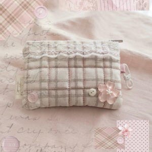 May include: A quilted fabric pouch featuring a light pink and beige plaid pattern. The pouch is accented with a white lace trim along the top, a small white button, and a translucent pink flower-shaped embellishment. A white plastic clip is attached.