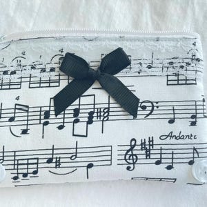 May include: A white zippered pouch with a musical note pattern, a black bow, and white lace trim. The pouch has two white buttons and the word "Andante" is printed on the fabric. The pouch is approximately 6 inches wide and 4 inches tall.