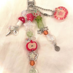 May include: A whimsical keychain featuring a variety of colorful charms. The charms include a red mushroom, a star, a strawberry, a green vegetable, and a red apple. The keychain is attached to a silver clasp and chain.