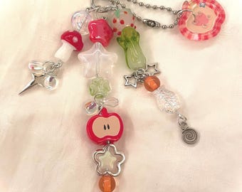 May include: A whimsical keychain featuring a variety of colorful charms. The charms include a red mushroom, a star, a strawberry, a green vegetable, and a red apple. The keychain is attached to a silver clasp and chain.