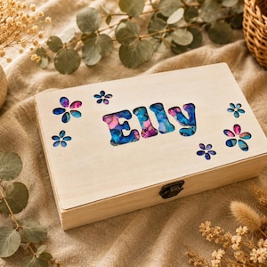 May include: A rectangular light-coloured wooden box featuring a cutout of the name ELLY and several flower-shaped cutouts on the lid. The cutouts are filled with a vibrant, multi-coloured resin inlay displaying shades of blue, pink, and purple.