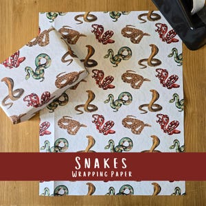 May include: White wrapping paper featuring a repeating pattern of watercolour snakes in brown, red, and green. A wrapped gift box is on the left. The text "SNAKES WRAPPING PAPER" is at the bottom.