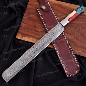 May include: A long, patterned steel chef's knife with a handle featuring red, white, and blue accents. The knife rests in a brown leather sheath with white stitching, on a wooden cutting board. The blade has a unique, swirling pattern.