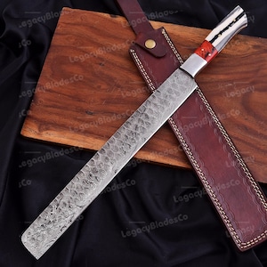 May include: A long, straight-edged knife with a patterned Damascus steel blade. The handle features a white bone-like section with a dark center line and a bright red accent piece. It is accompanied by a brown leather sheath with stitching.