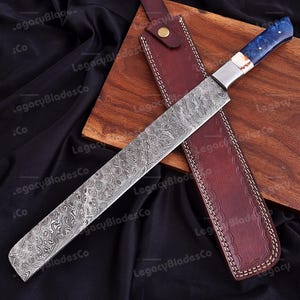 May include: A long, Damascus steel knife with a blue and white handle, resting on a wooden cutting board. The knife is accompanied by a brown leather sheath with white stitching. The blade has a unique, patterned design.