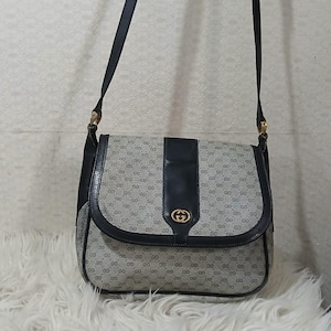 May include: A vintage shoulder bag with a repeating pattern of interlocking "G" logos. The bag features a flap closure with a gold-tone emblem and a black leather strap. The bag is set against a neutral background.