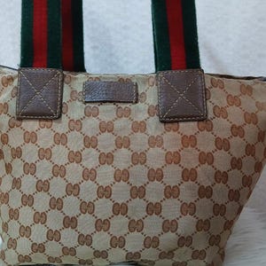 May include: A brown and beige tote bag with a green and red striped handle. The bag is made of a canvas material with a repeating pattern of the Gucci logo.