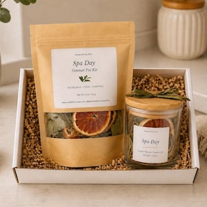 May include: A white gift box containing a brown paper pouch labeled Spa Day Simmer Pot Kit with dried citrus and eucalyptus, and a glass jar with a wooden lid and green ribbon, also labeled Spa Day, filled with dried botanicals and citrus slices.