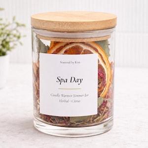May include: A clear glass jar with a light wood lid filled with dried orange slices, herbs, and botanicals. The white label reads: Seasonal by Kim, Spa Day, Candle Warmer Simmer Jar, Herbal Citrus. The jar is cylindrical with a flat base.