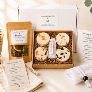 May include: A Seasonal by Kim Fall botanical collection gift box containing four round shower steamers with dried floral and spice toppings, a pouch of Harvest botanical tea, a scent guide card, a recipe card, and a white mesh drawstring bag.