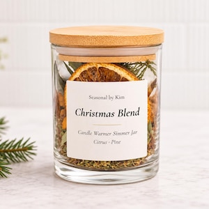 May include: A clear glass jar with a light wood lid containing a Christmas Blend simmer pot mixture. The white label reads Seasonal by Kim, Christmas Blend, Candle Warmer Simmer Jar, Citrus Pine. Dried orange slices and greenery are visible.
