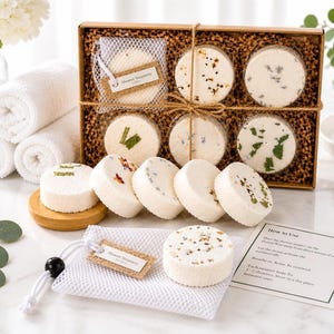 May include: A set of round white shower steamers with botanical inclusions, packaged in a brown cardboard box with shredded paper filler. Includes a white mesh drawstring bag, a wooden base, and an instructional card labeled How to Use.