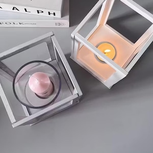 May include: Two white geometric candle holders with glass panels. One holds a lit orange candle, the other a pink candle in a clear glass. Books with the text "ALPH" and "FOLKHOME" are in the background.