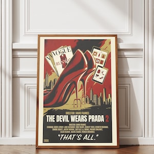May include: A framed poster with a brown wooden frame. The poster features a stylized image of a red high-heeled shoe, a Vogue magazine cover, and the text "THE DEVIL WEARS PRADA 2". The poster is set against a white wall.
