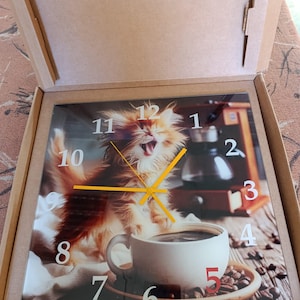 May include: Square wall clock featuring a fluffy orange and white cat yawning, with a coffee cup and coffee beans. The clock face has white numbers and yellow hands. The clock is in a cardboard box.