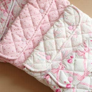 May include: Three quilted fabric pouches featuring pink and white patterns. The left pouch has a pink ribbon design, the centre has a pink and white striped pattern with small dots, and the right has a pink rose and ribbon floral print.