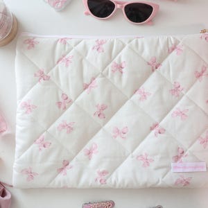 May include: A white quilted laptop sleeve with a pink floral pattern. The sleeve is rectangular with a diagonal stitched pattern and a gold zip. Other items in the image include sunglasses, perfume, and makeup.