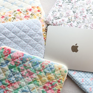 May include: A collection of quilted fabric laptop sleeves featuring various patterns including floral prints, blue gingham, and light blue bows, arranged around a silver laptop with a visible Apple logo on the lid. Each sleeve has a diamond-stitched texture.