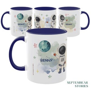 Könnte beinhalten: Ein weißer Keramikbecher mit dunkelblauem Henkel und Innenraum, verziert mit einer Aquarellillustration eines Astronauten, eines Planeten und Sternen. Der Name BENNY ist in dunkelblauer Schrift gedruckt, und der Markenname SEPTEMBEAR STORIES ist sichtbar.