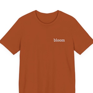 May include: A rust-colored t-shirt with short sleeves. The word "bloom" is printed in white on the upper chest area. The shirt is made of a soft material and is suitable for casual wear.