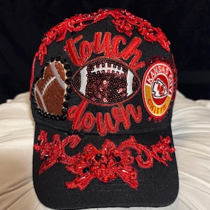 May include: Black baseball cap with red embellishments and the words "Touch Down" in red script. Features a football and Kansas City Chiefs logo. The cap is decorated with red sequins and beadwork.