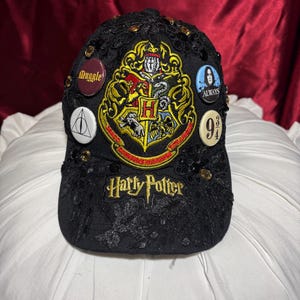 May include: Black baseball cap with lace detailing and gold accents. Features a large embroidered Hogwarts crest and the words "Harry Potter." Includes several pin-on buttons with text and symbols from the Harry Potter series.