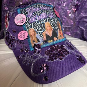 May include: Purple baseball cap with sequin and bead embellishments. The cap features a patch with the text "You can't sit with us!" and images of characters. Additional pins include "Get in loser" and "on Wednesdays we wear pink."